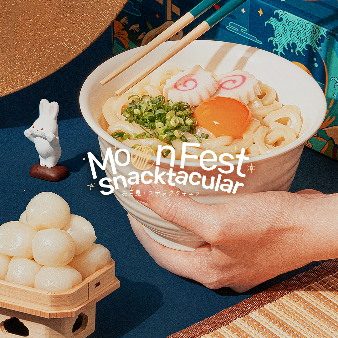 A pair of hands holds a bowl of Tsukimi udon, surrounded by Tsukimi Moon Festival decorations.