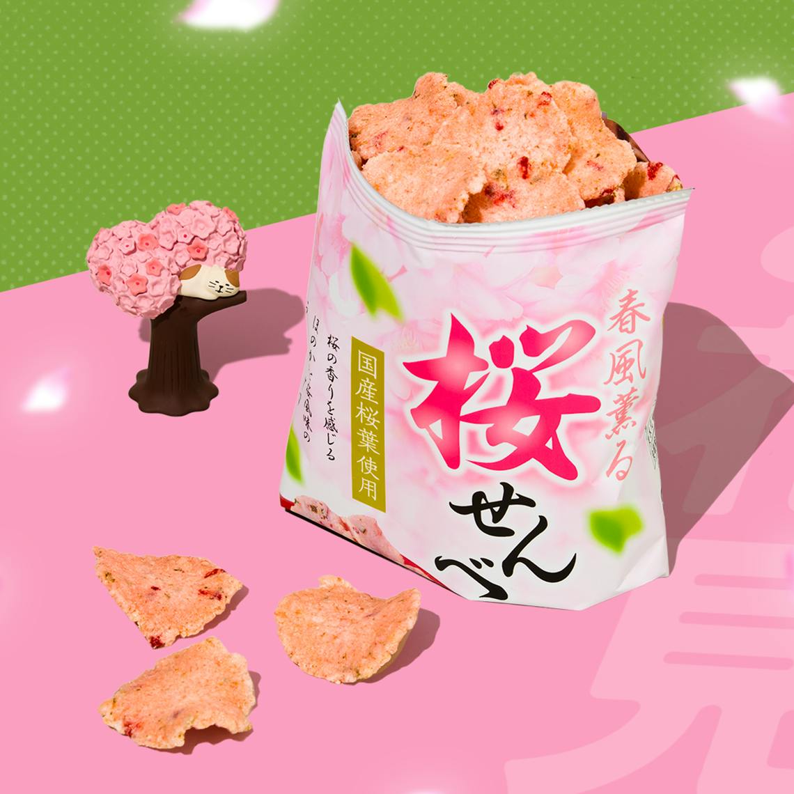 Bag of sakura shrimp rice crackers on pink background with figurine of cat resting on cherry blossom tree branch next to it