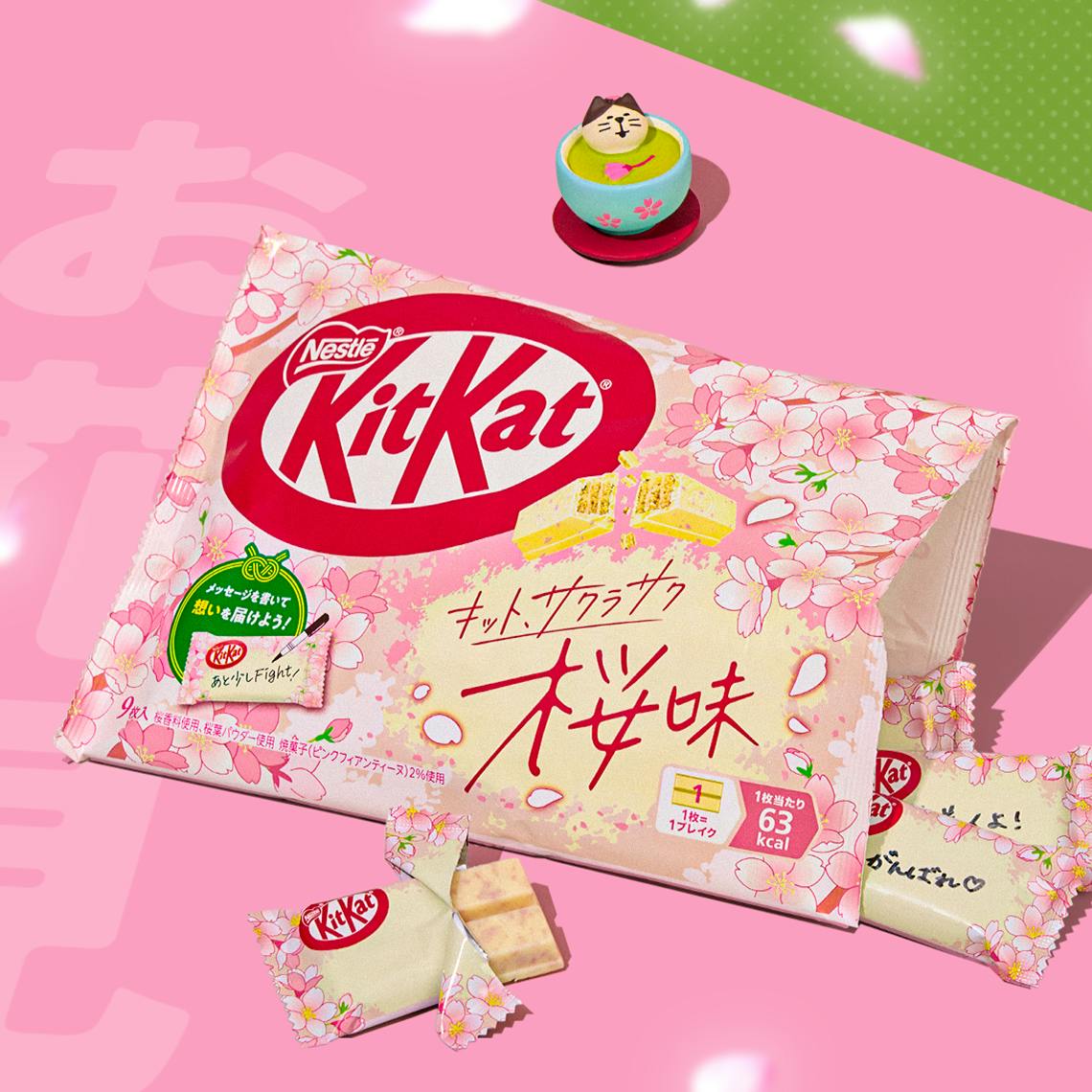 Package of sakura KitKats on pink background with figurine of cat resting in a cup of matcha in the back